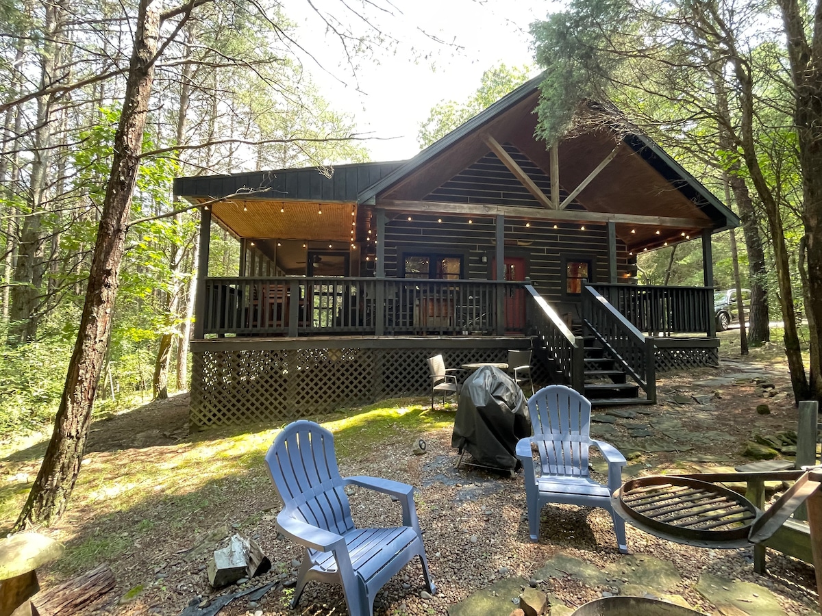 A spacious cabin is nestled among trees, featuring a large porch with seating. Two adirondack chairs are positioned on a gravel area in front of the cabin. A fire pit is visible nearby, surrounded by a natural landscape.