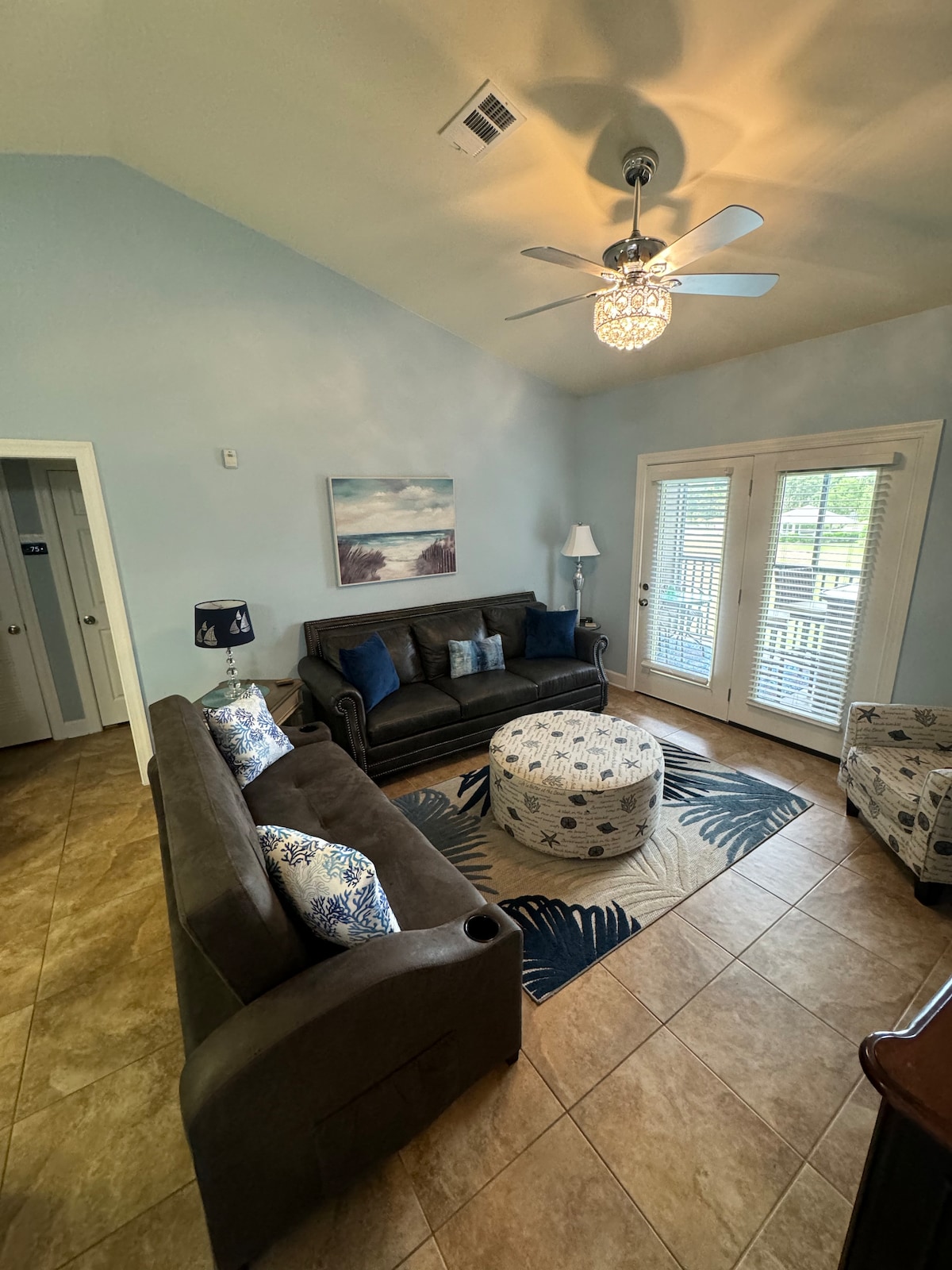 A cozy living area features a dark sofa and a light armchair, accompanied by a patterned ottoman. A ceiling fan hangs above, and natural light enters through glass doors that open onto a screened balcony. Soft blue walls enhance the inviting atmosphere.
