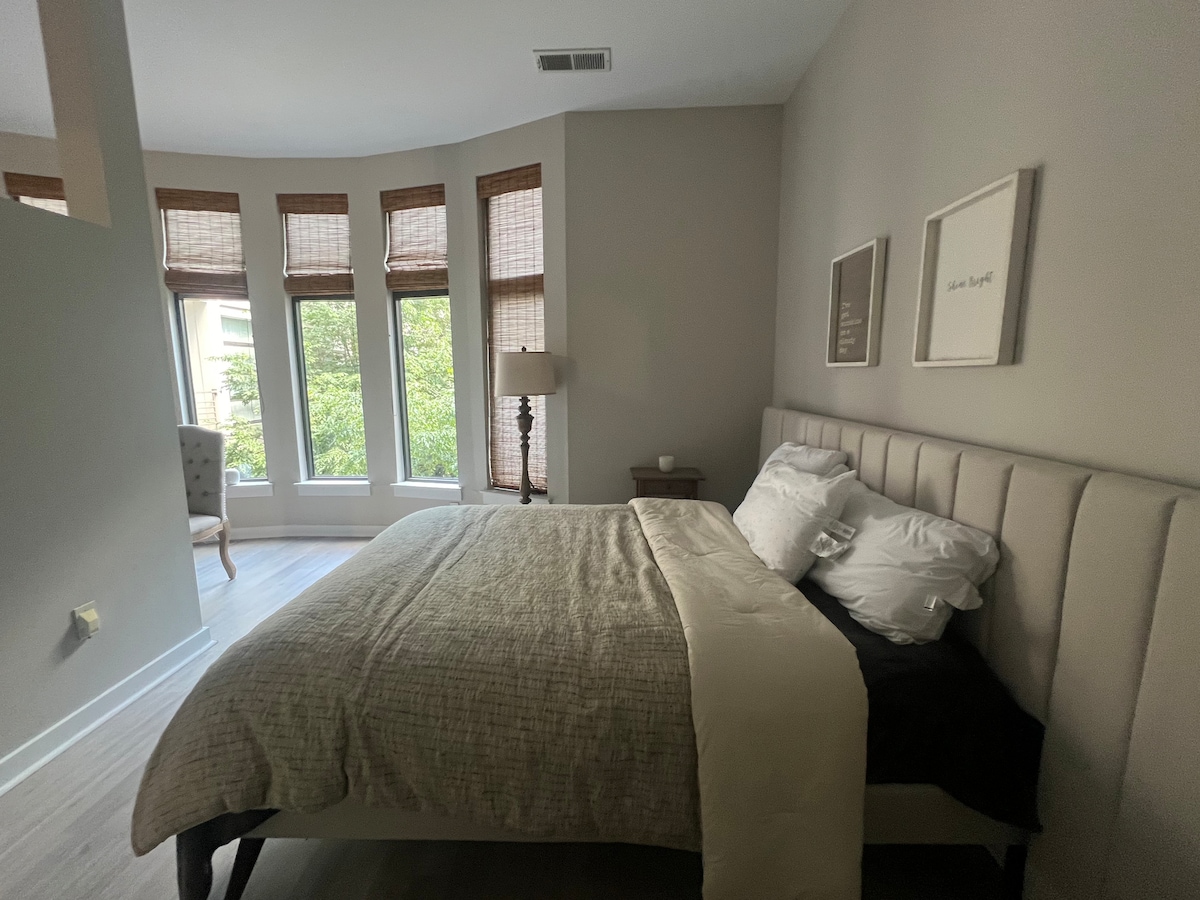 A serene bedroom is highlighted by a spacious bed dressed in neutral bedding. Natural light filters through multiple large windows adorned with bamboo shades, illuminating the soft color palette. A bedside table holds a lamp, while a comfortable chair is placed in the corner.