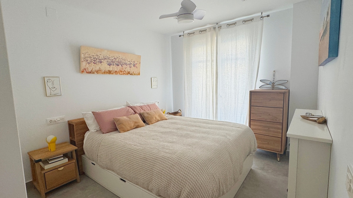 A light-filled bedroom features a spacious bed with neutral bedding and decorative pillows. Natural light filters through sheer curtains, illuminating the area. A wooden nightstand and dresser provide storage, while wall art adds a touch of character to the softly colored walls.