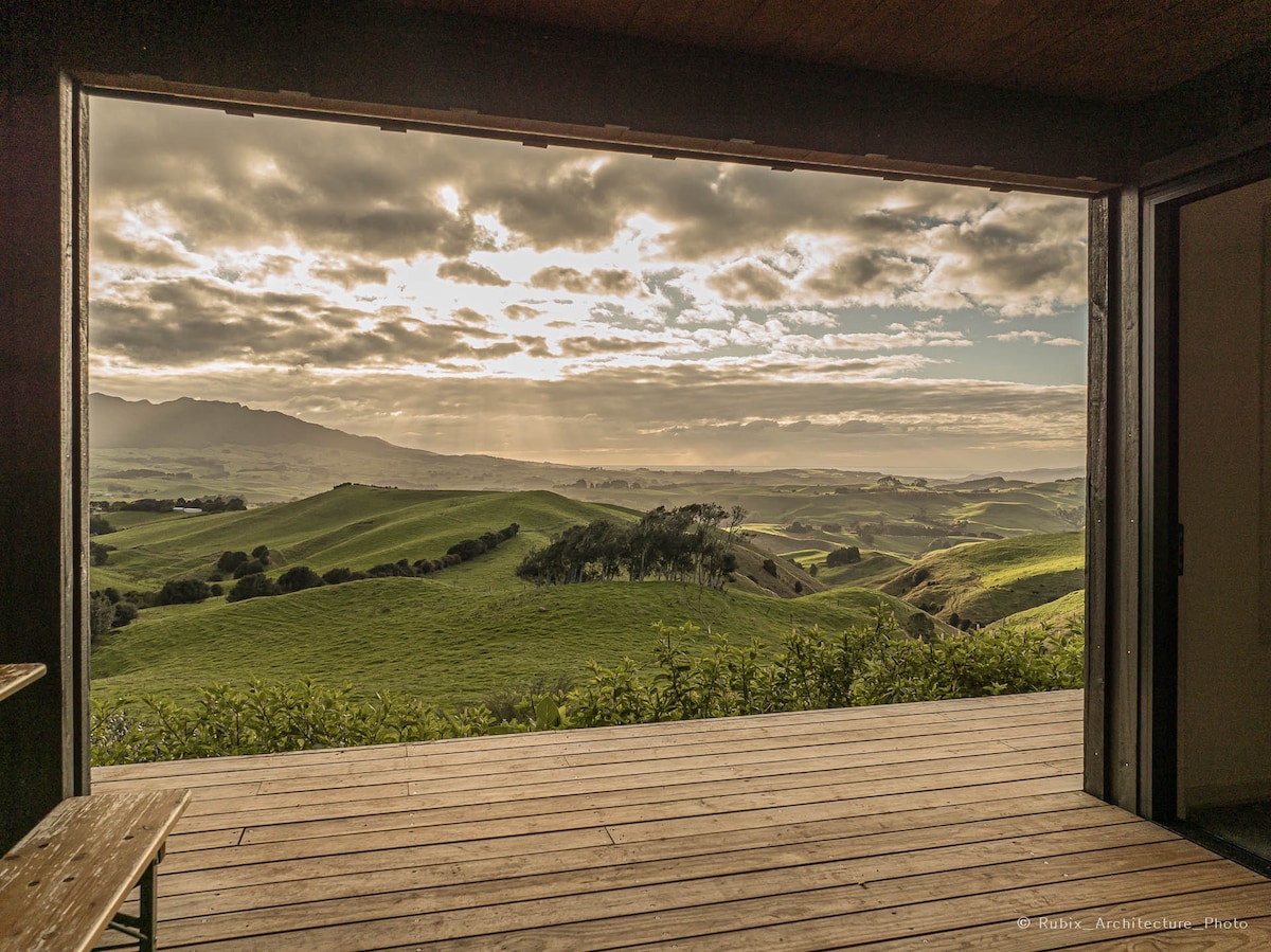 A wide opening reveals a breathtaking view of rolling green hills under a dramatic sky. The natural landscape is illuminated by soft rays of sunlight filtering through clouds, creating a peaceful ambiance. The wooden flooring leads to the outdoor space, enhancing the connection with nature.