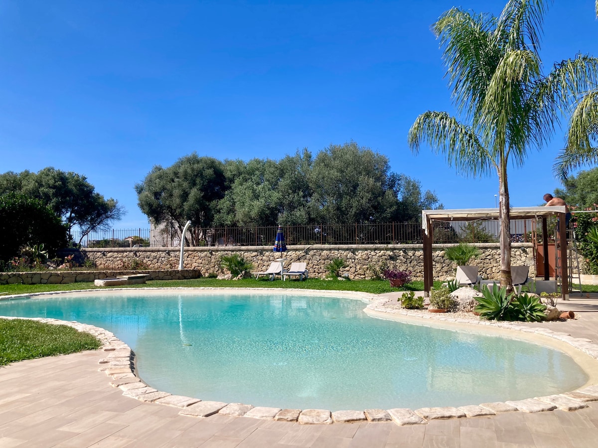 A serene pool area is surrounded by lush greenery and neatly arranged landscaping. Sun loungers are positioned nearby, offering relaxation opportunities under the clear blue sky. The pool features a gentle curve and is complemented by palm trees that enhance the tranquil setting.