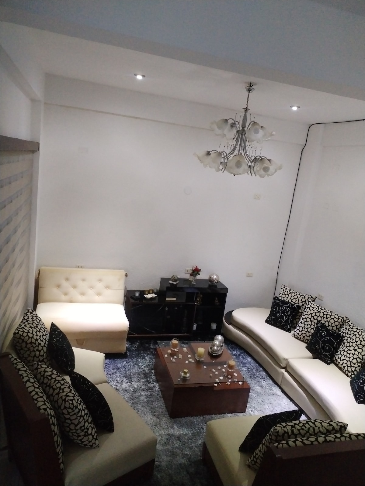 A cozy living area is furnished with two white sofas and a brown wooden sofa, all adorned with patterned cushions. A wooden coffee table occupies the center, surrounded by decorative items and candles. A modern chandelier with white shades hangs from the ceiling, providing soft lighting.