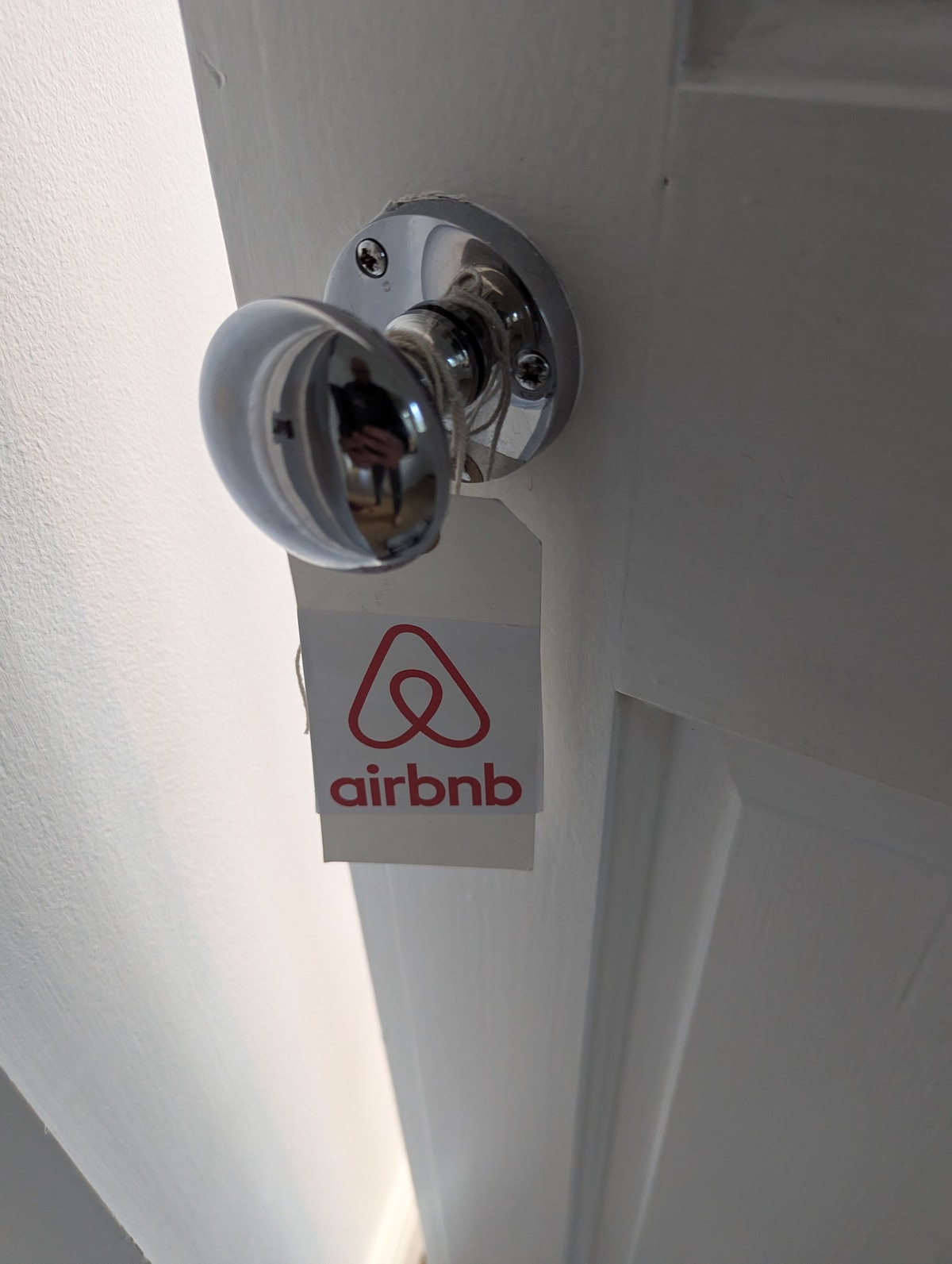 A close-up view of a polished doorknob is presented, featuring a clear tag with the Airbnb logo attached. The smooth surface of the door reflects natural light, highlighting the clean lines of the doorframe and the minimalist aesthetic.