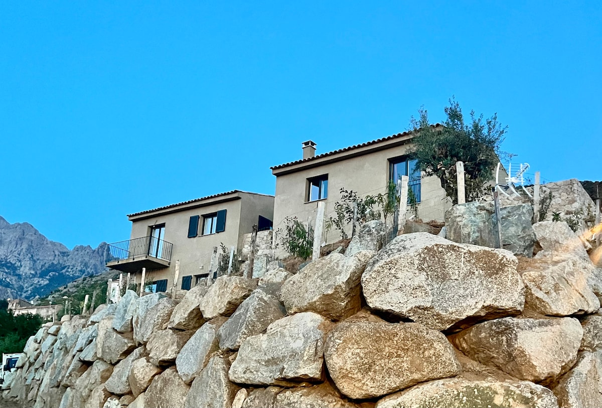 Two elegant houses are perched on a hillside, surrounded by rocky boulders and a backdrop of mountains. Large windows offer views of the landscape, while a terrace extends from the first floor, providing an outdoor space to enjoy the scenery.