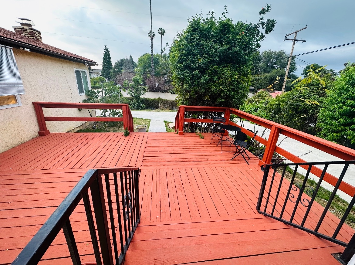 A spacious wooden deck features a vibrant red finish, providing an outdoor seating area with a small table and chairs. Lush greenery and palm trees are visible in the background, while a pathway extends towards the street, enhancing the serene atmosphere.