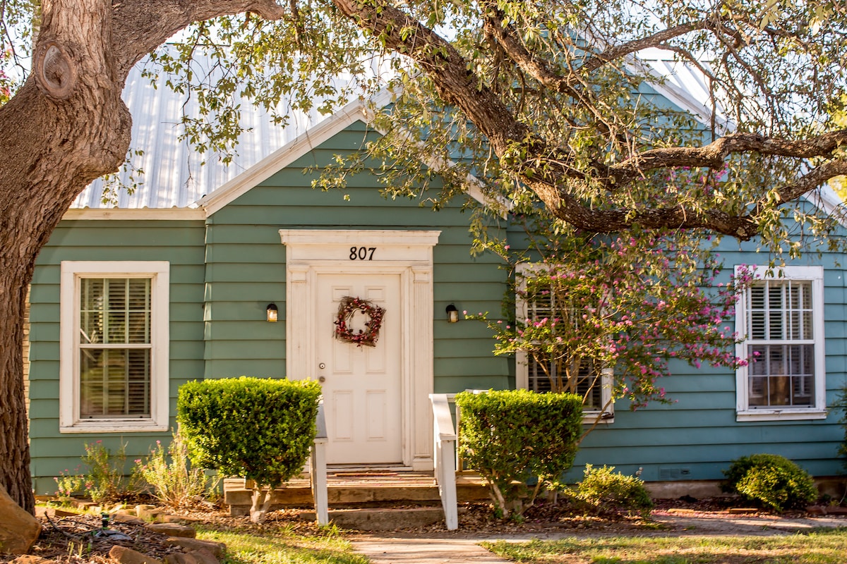A charming green house features a welcoming front door adorned with a wreath. Two manicured bushes frame the entrance, and vibrant flowers are visible by the steps. A large tree provides shade, enhancing the inviting exterior.