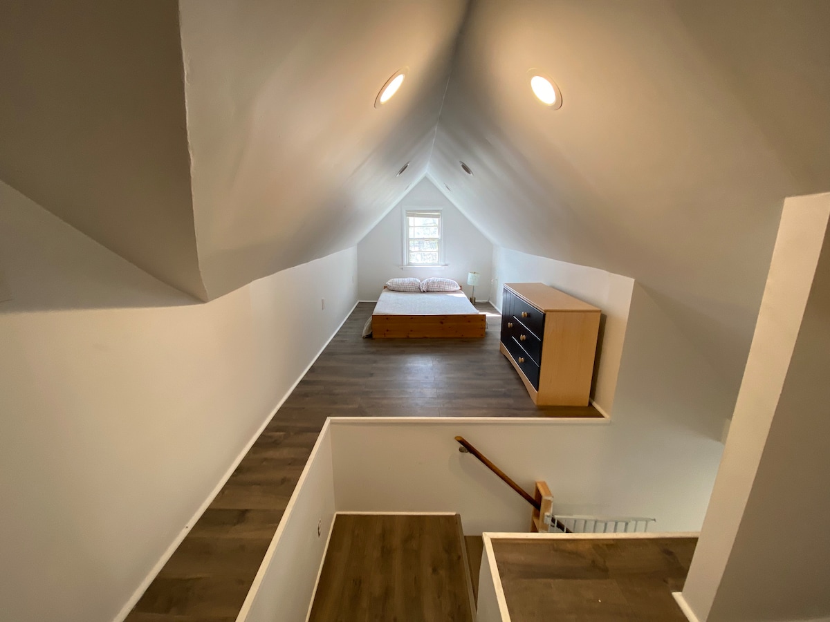 An airy loft space is captured from an elevated angle, featuring a spacious bed positioned under a high, sloped ceiling. A wooden dresser with multiple drawers is located against one wall, while a staircase provides access to the area.