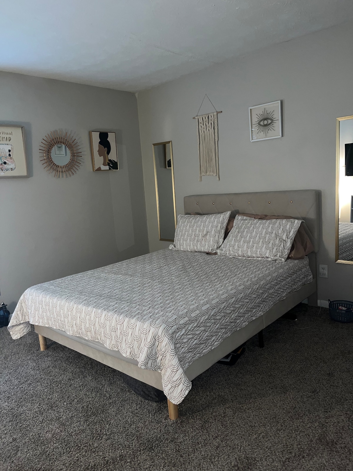 A comfortable bedroom features a spacious bed with a patterned duvet and two pillows. A wall mirror reflects the room's décor, which includes wall art and a decorative sunburst mirror. Soft carpeting covers the floor, contributing to a cozy atmosphere.