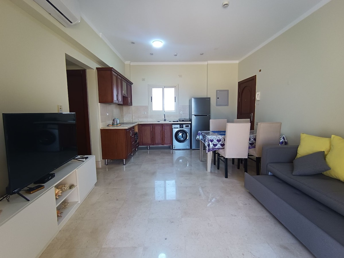 A bright and airy living space features a modern kitchen with wooden cabinets and stainless-steel appliances. A dining table with four chairs is positioned next to the kitchen. A couch with cushions adds comfort, while a large window allows natural light to fill the room.