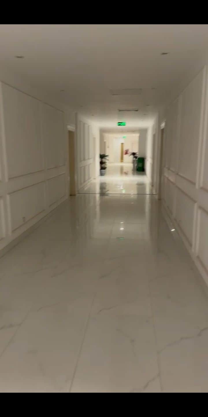 A long hallway is shown, featuring sleek, polished marble floors that reflect the soft overhead lighting. White walls are embellished with classic paneling, and doors are visible along the corridor. At the far end, light spills from a doorway into a bright space.