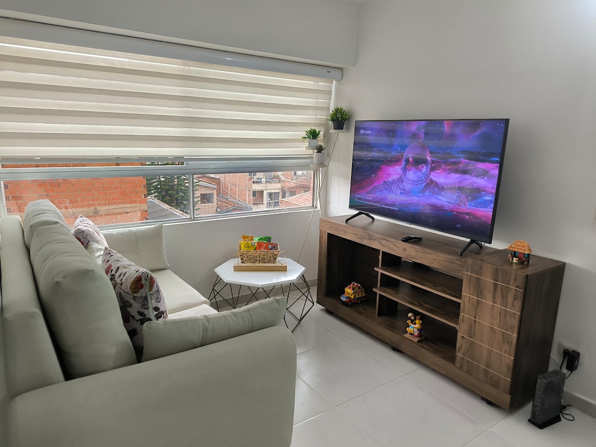 A comfortable living area features a light-colored sofa adorned with decorative cushions. A small, modern coffee table is positioned in front of the sofa. A large television displays a colorful image, while natural light fills the space through window coverings.