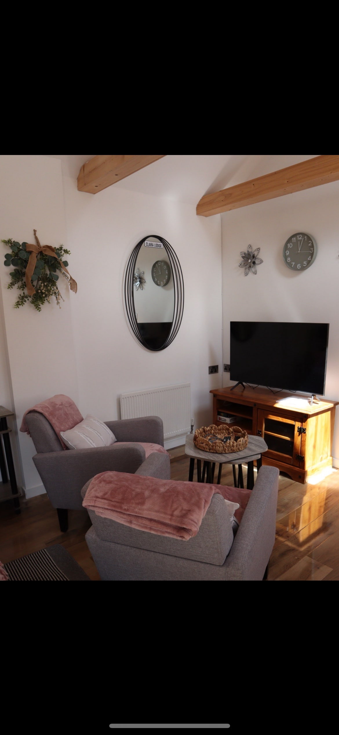 A cozy living area features two gray armchairs with soft pink throws. A wooden coffee table holds decorative items, while a flat-screen television is mounted on the wall. Light wood beams highlight the ceiling, and a large oval mirror reflects the room's warmth.