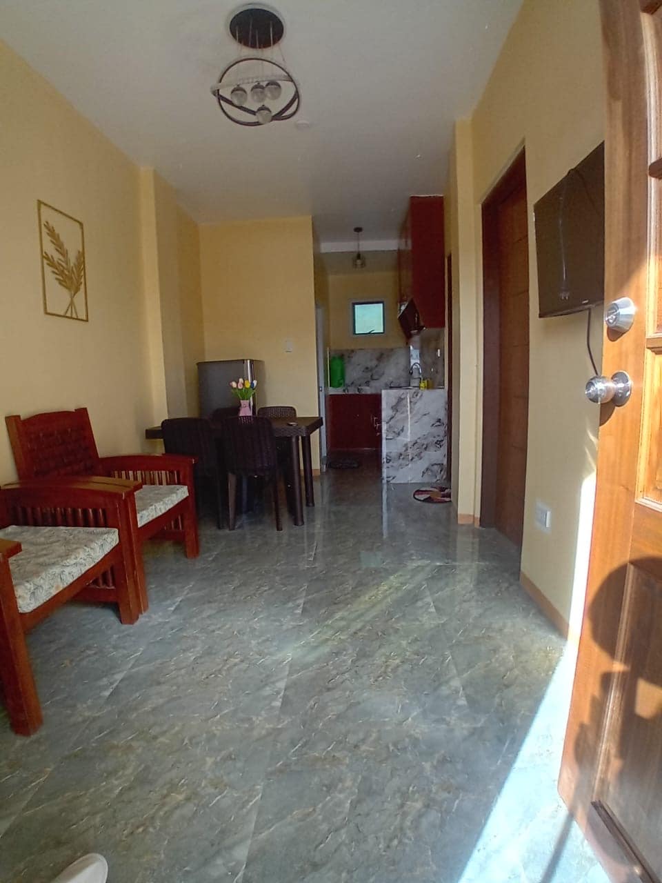 A spacious hallway leads into an inviting living area, featuring wooden chairs and a dining table for four. The kitchenette is visible in the background, showcasing modern appliances against a marble countertop. Natural light filters in, enhancing the warm tones of the room.