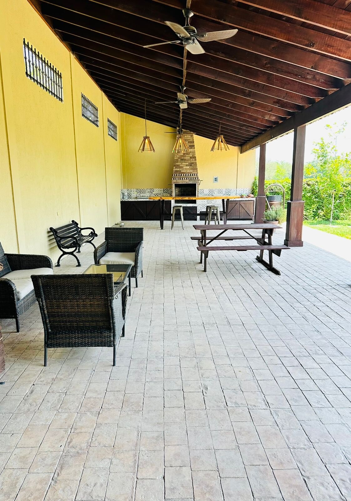 An outdoor social area is presented with a spacious covered patio featuring wicker chairs and a long picnic table. A built-in grill is located in the background, accompanied by decorative lighting. Surrounding greenery enhances the natural ambiance of the space.