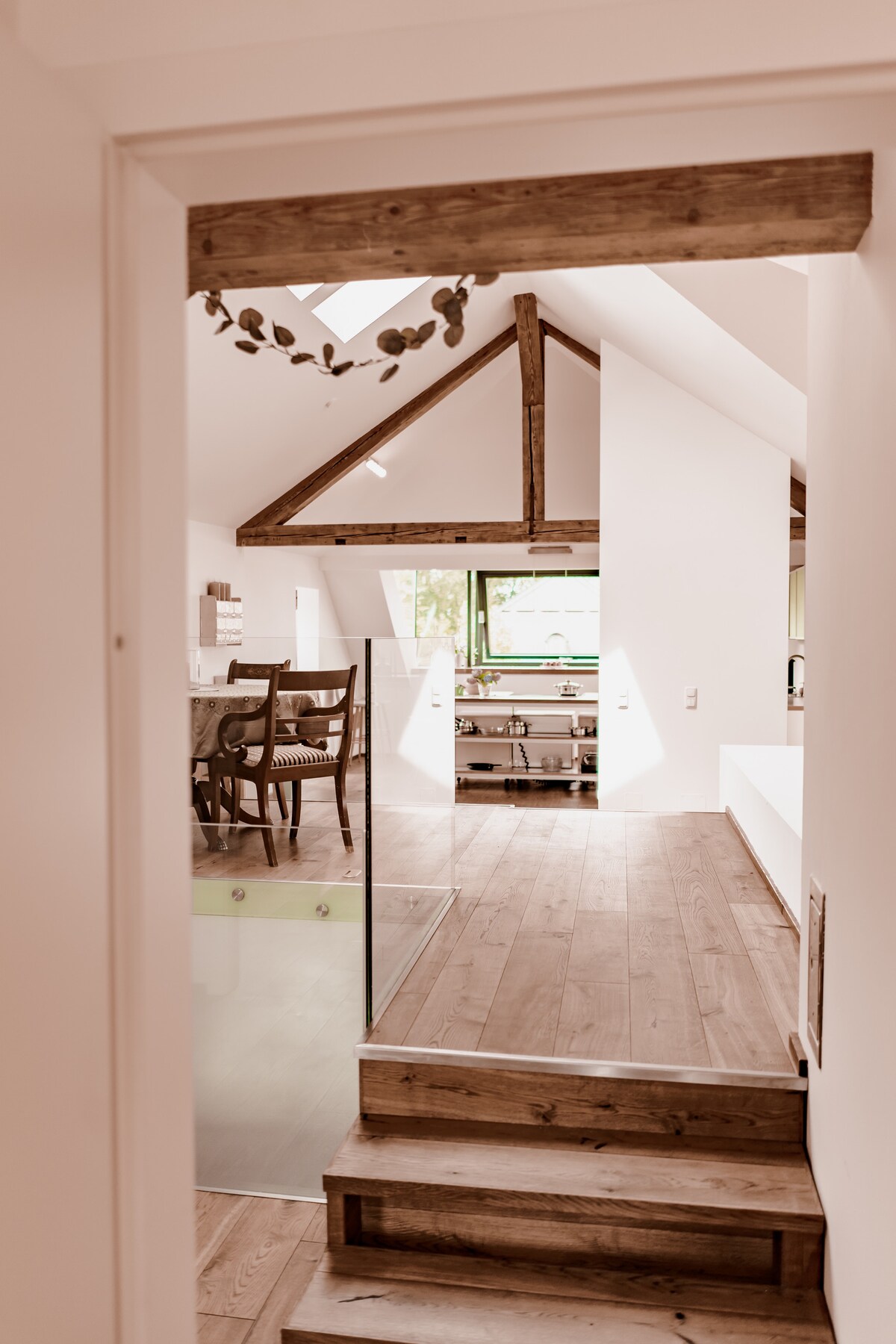 A sunlit entryway features wooden steps leading into an open living area. Exposed beams add character to the ceiling, while large windows bring in natural light. A dining table with chairs is visible, located near a modern kitchen design.