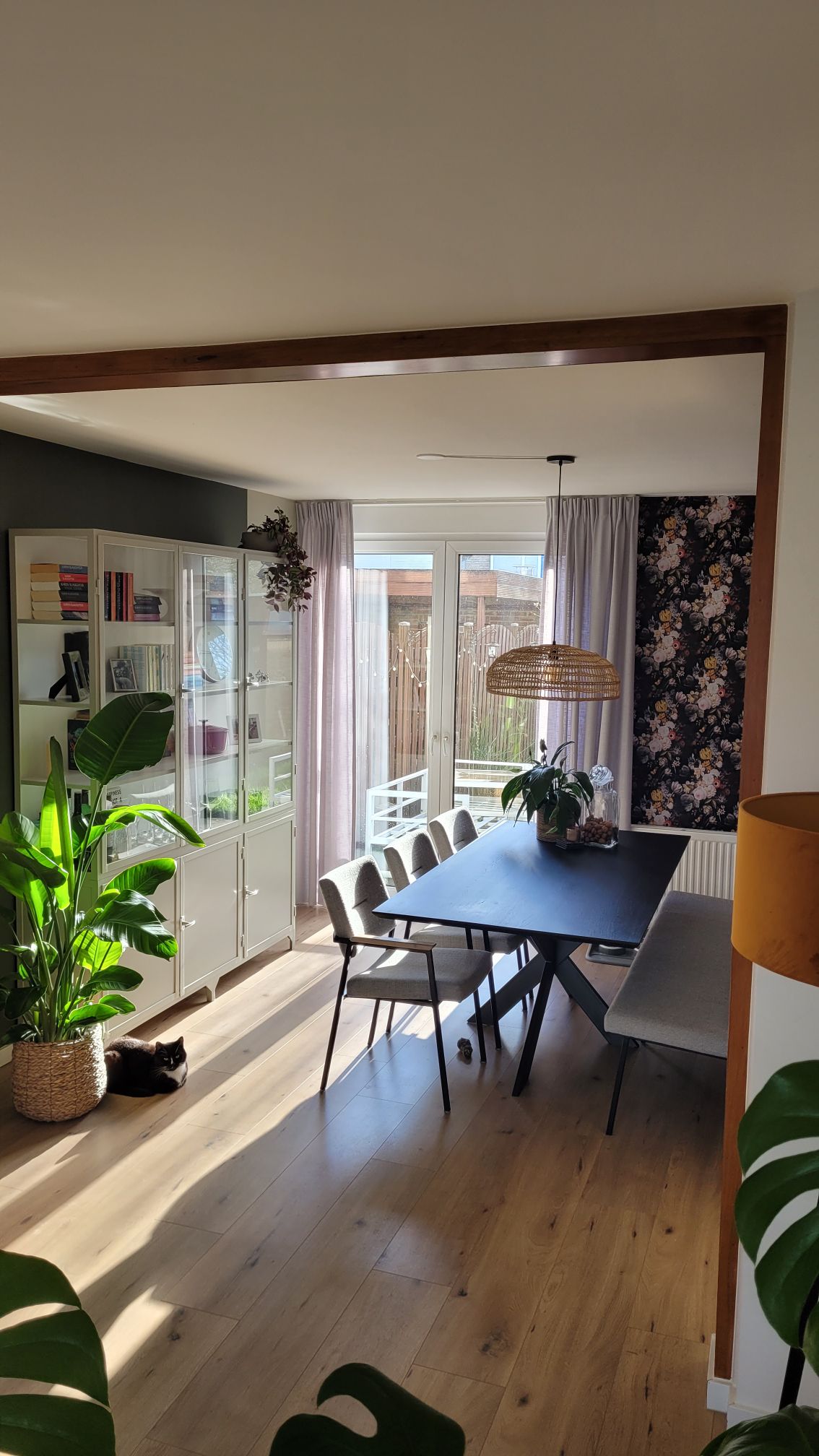 A dining area is presented with a sleek black table surrounded by five modern chairs. Large windows allow natural light to fill the space, complemented by indoor plants and a decorative cabinet. The walls feature a floral accent, enhancing the room's layout.