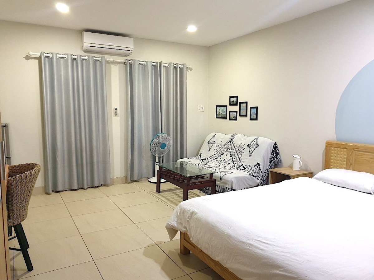 A bright room featuring a spacious bed and a seating area with a wicker chair and a sofa adorned with patterned cushions. A fan is positioned nearby, and light is provided by overhead fixtures. Light-colored curtains frame the windows, enhancing the airy ambience.