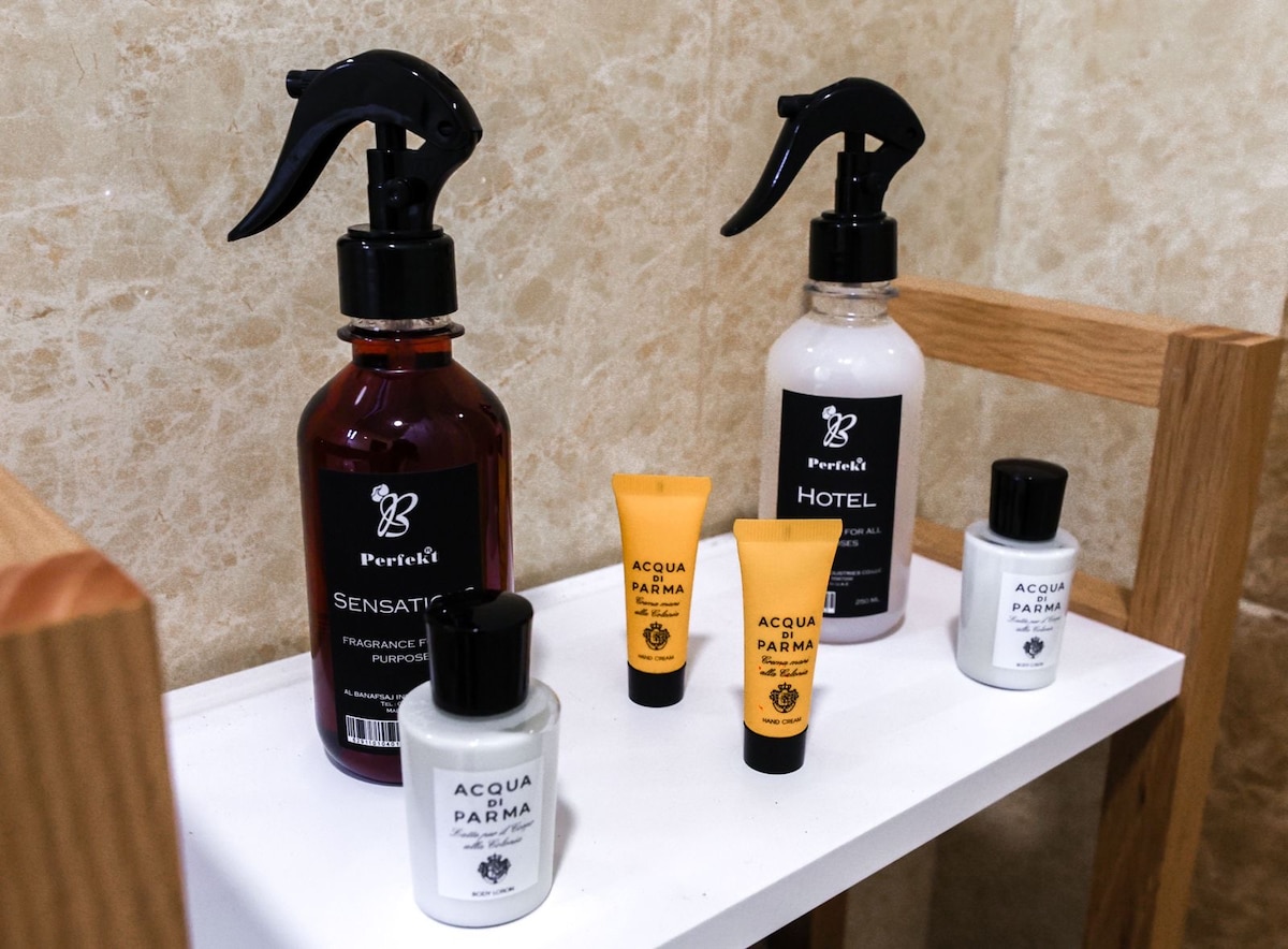 A selection of personal care items is arranged on a wooden shelf in the bathroom. Two large spray bottles labeled 'Sensation' and 'Hotel' sit next to three travel-sized tubes of shampoo, conditioner, and lotion, offering guests convenient toiletries.