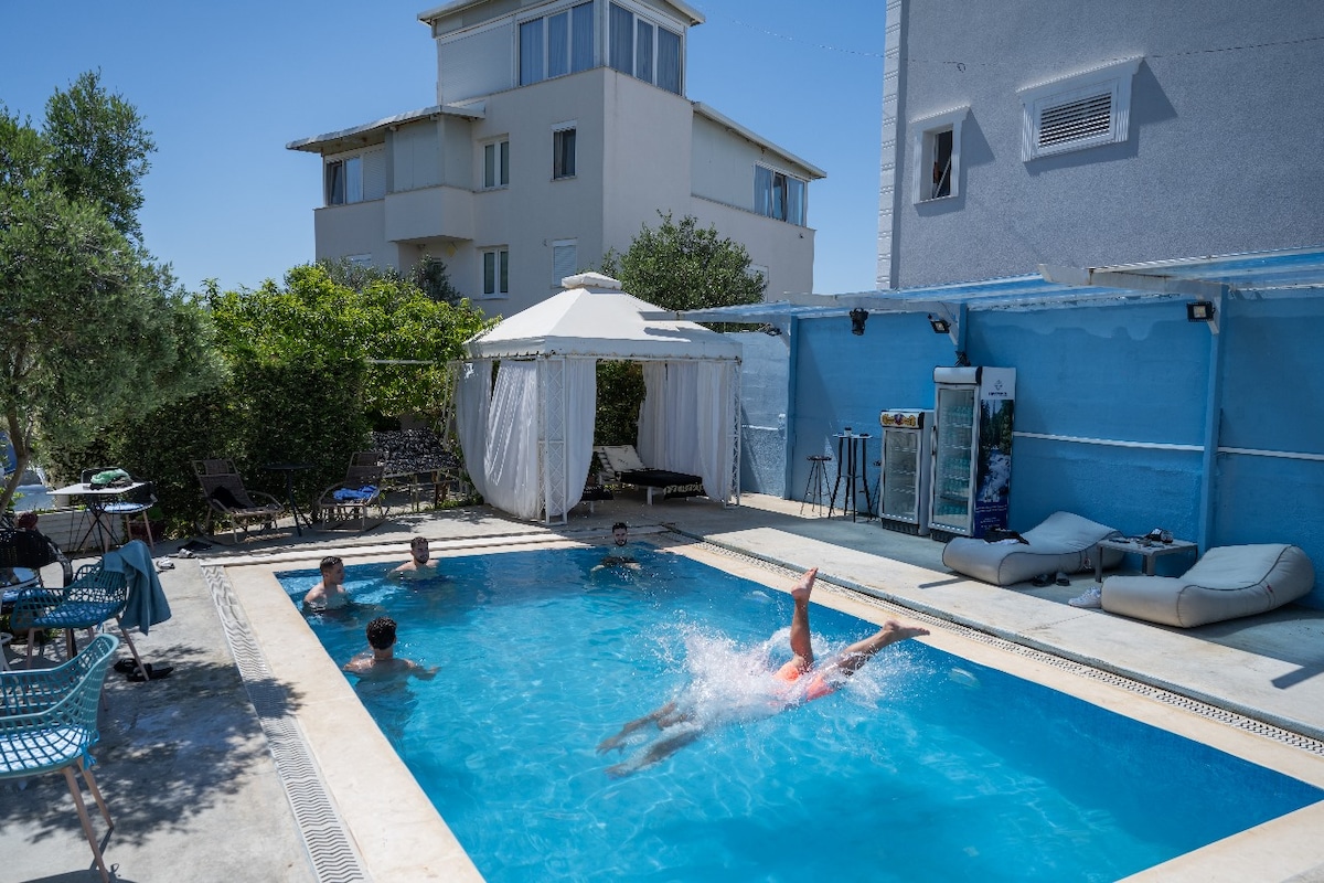 A private outdoor pool area is shown, featuring clear blue water and a shaded gazebo in the background. Loungers are positioned around the pool, where individuals are seen enjoying the water. Surrounding greenery enhances the outdoor setting.