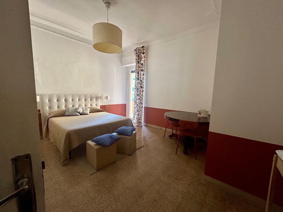 A spacious bedroom is highlighted by a large bed with a tufted headboard, accompanied by decorative pillows. Natural light enters through a window with vibrant patterned curtains. A small table and chairs are visible, creating a functional area for relaxation or work.
