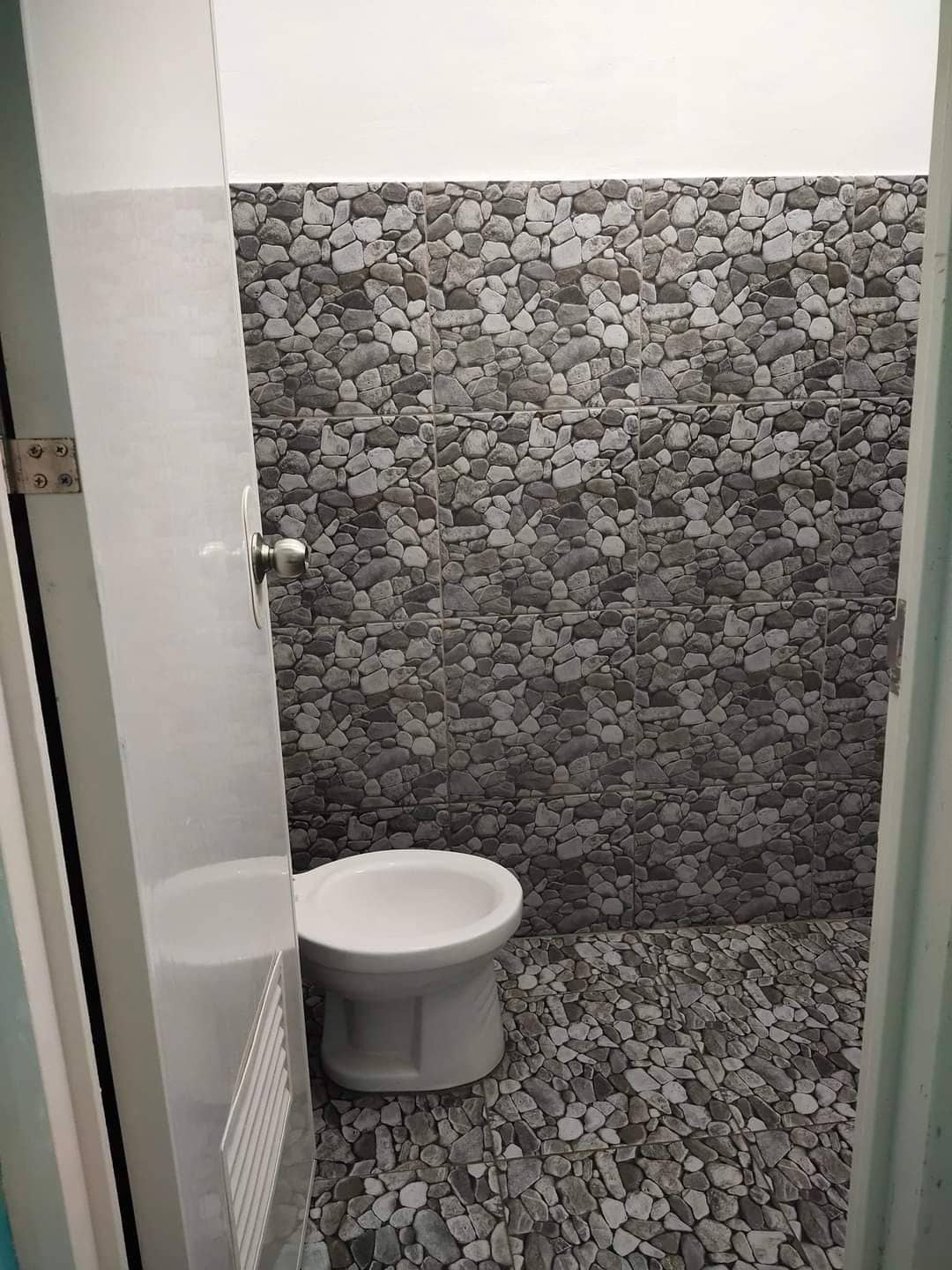 A compact bathroom features a white toilet positioned against a wall adorned with textured stone tiles, extending from the floor to the ceiling. Natural light illuminates the space, enhancing the earthy tones of the tiles.