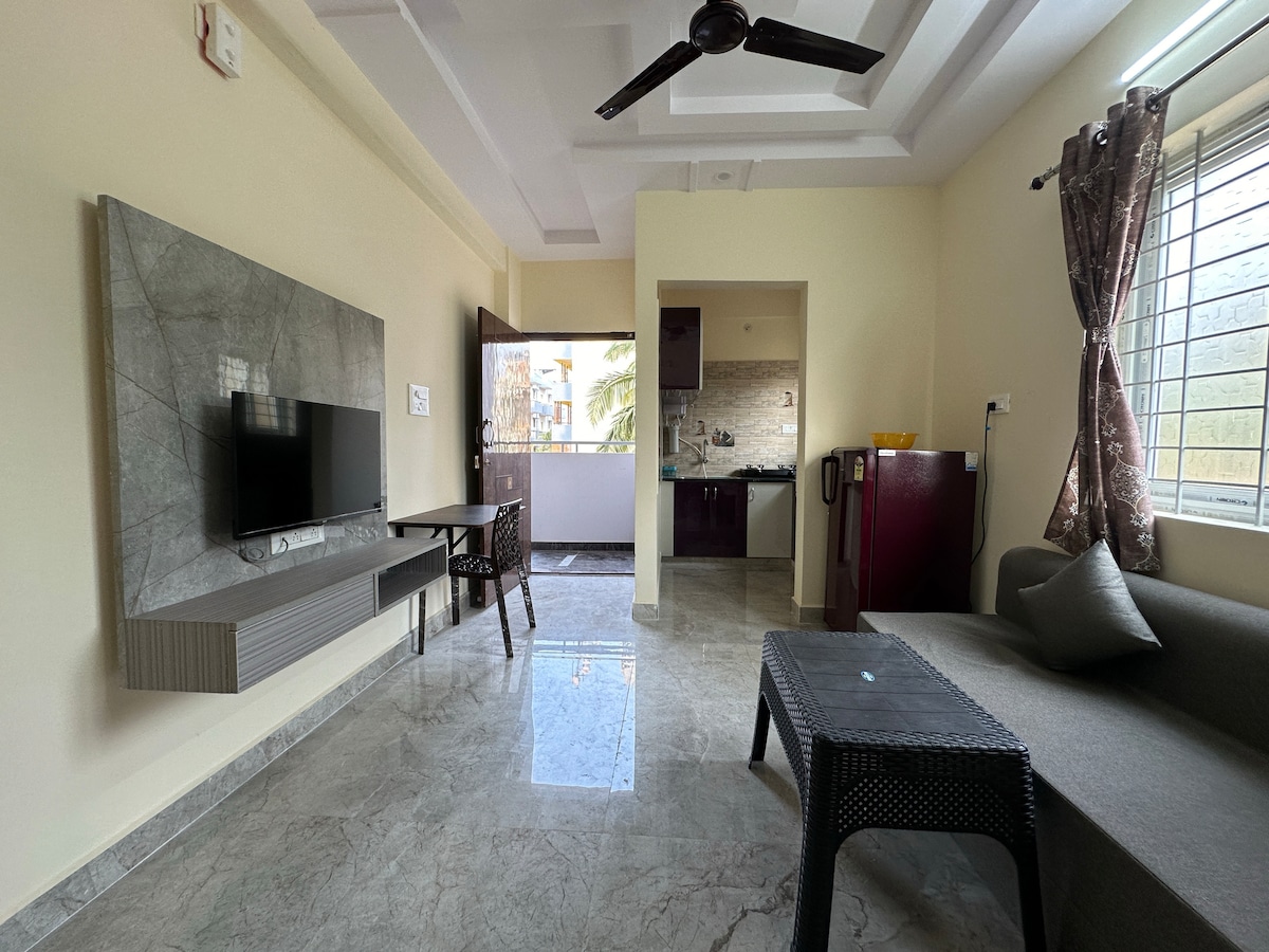An inviting living area is presented with a sleek marble floor and neutral-toned walls. A modern television is mounted on a decorative wall unit, while a grey sofa offers a comfortable seating option. Natural light enters through a window that opens to the exterior.