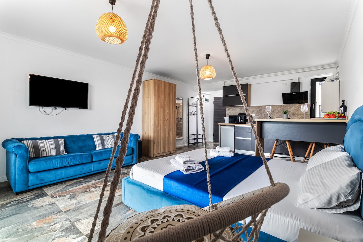 A modern studio space showcases a comfortable double bed with a blue and white bedding set. A cozy blue sofa and a hanging chair add to the seating options. An open kitchenette features dark cabinetry, and a dining table is set for four. Large windows allow for ample natural light.