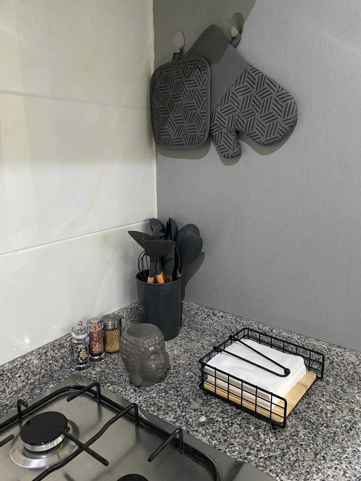 A modern kitchen countertop features a collection of kitchen utensils organized in a holder beside a decorative Buddha statue. Hanging above, two grey oven mitts are displayed. A wire basket holds a stack of napkins, complementing the sleek grey wall and granite surface.