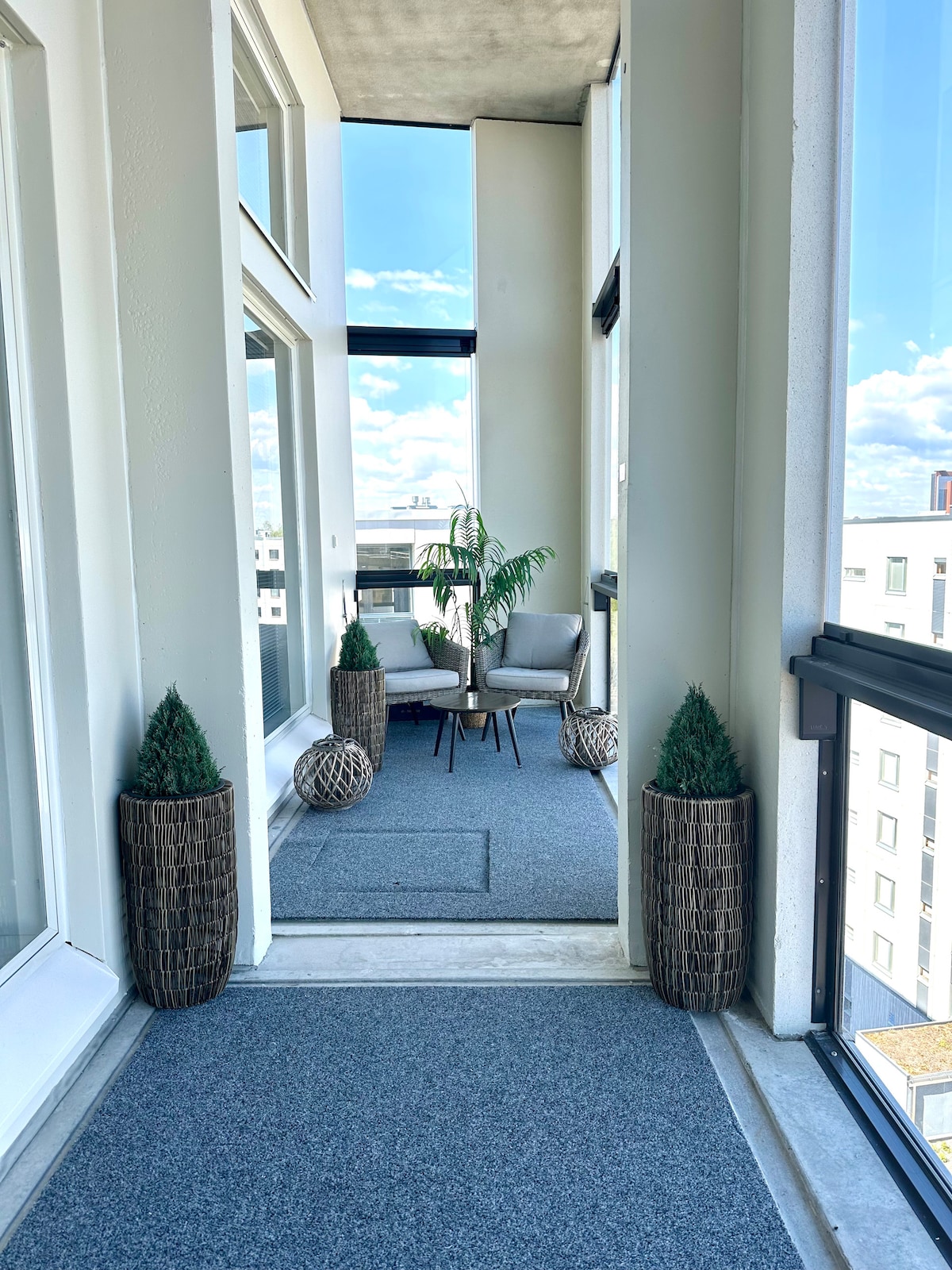 A hallway features large windows allowing natural light to fill the space. Two comfortable chairs are positioned beside a small table, while tall decorative planters with greenery are placed at each end of the area. The flooring is covered with a soft, neutral carpet.