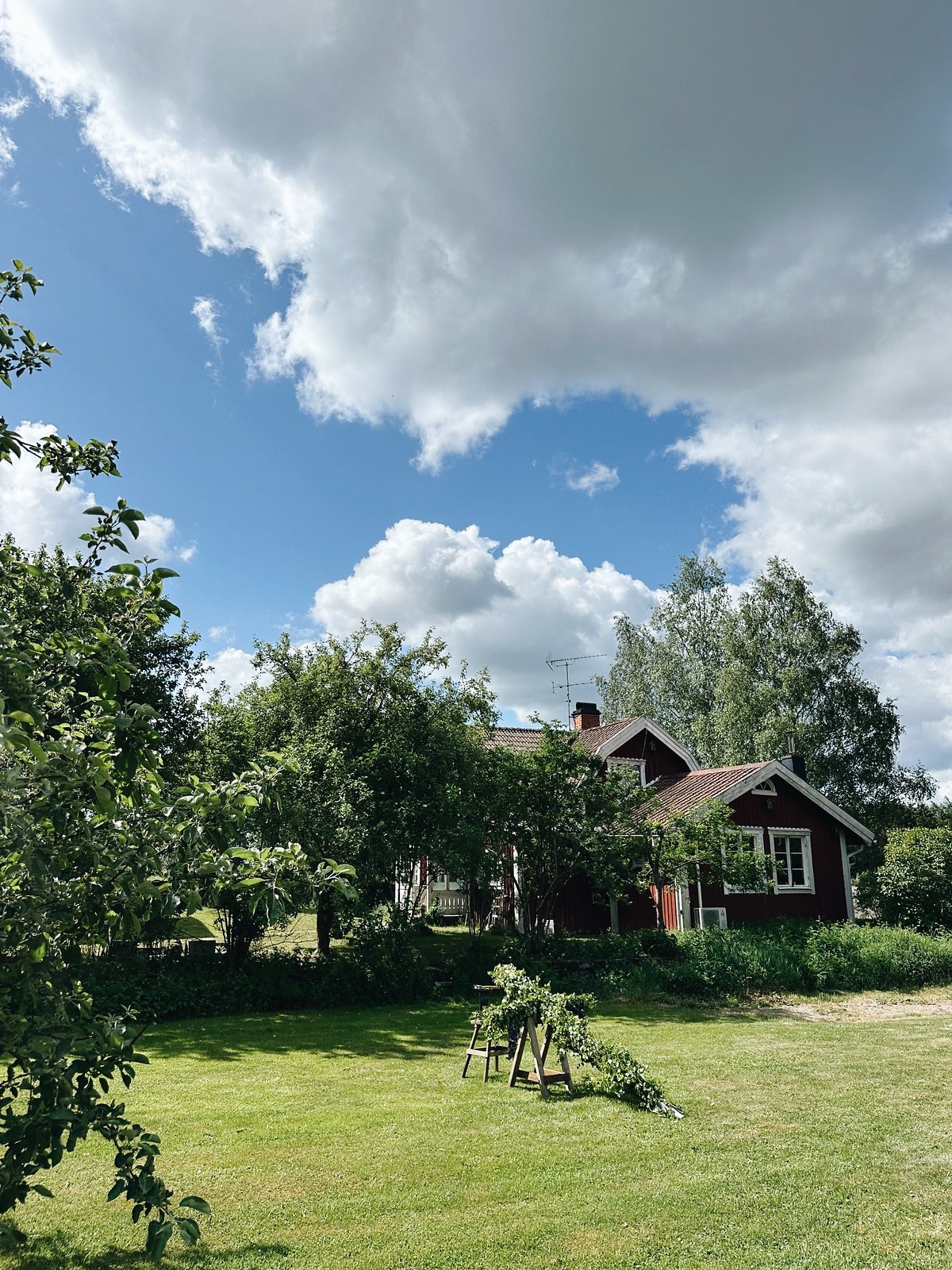 A traditional red house is set against a backdrop of lush greenery and a bright blue sky dotted with white clouds. Apple trees and other foliage surround the home, while a neatly maintained lawn offers open space for outdoor activities.