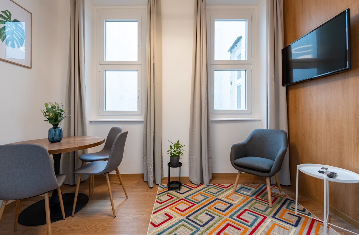 A cozy living area is highlighted by a round dining table surrounded by three grey chairs. A small plant sits on a black stand beside a comfortable armchair. Natural light fills the space through two windows, which are framed by soft curtains. A wall-mounted television adds to the modern decor.