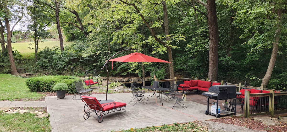 A spacious outdoor patio is surrounded by greenery, featuring comfortable red lounge chairs and a sectional couch. An umbrella provides shade over a dining table, while a grill sits in one corner. A tranquil stream can be seen in the backdrop.