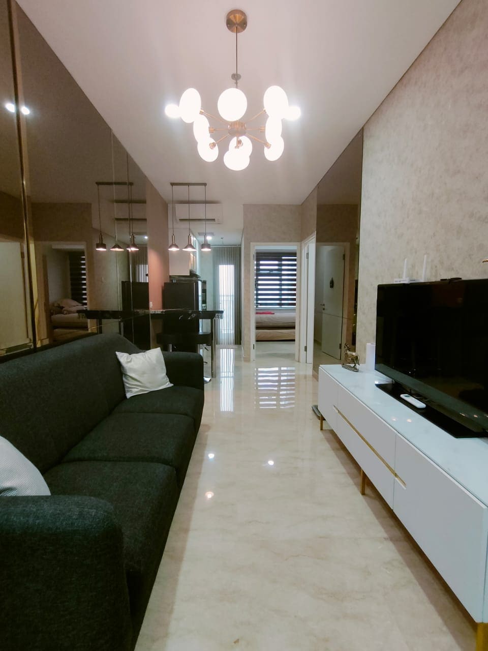 A modern living area features a dark gray sofa adorned with a white pillow. A contemporary light fixture hangs from the ceiling, illuminating the space. A flat-screen TV is positioned on a white console, while glass doors at the end lead to a balcony, enhancing natural light.