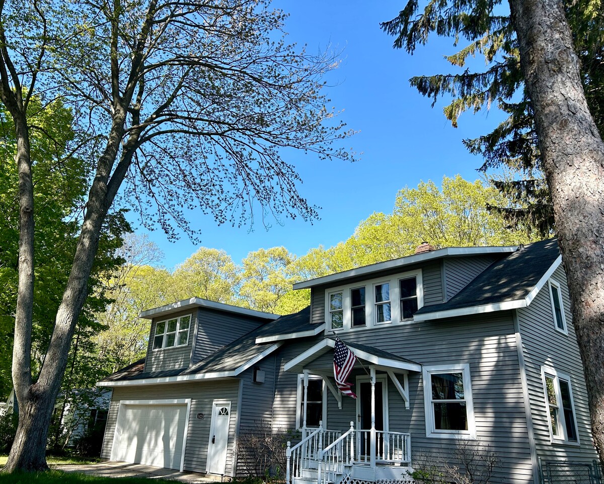 The exterior of the cottage is depicted, featuring a gray facade and a welcoming front porch with white railings. A flag is shown waving gently, while surrounding trees display fresh green foliage under a bright blue sky.