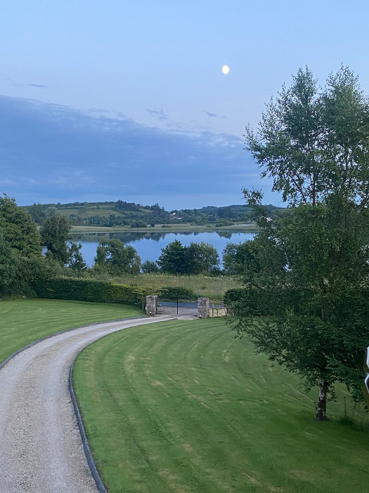 A gentle gravel driveway curves through well-manicured green lawns, leading to a gate. Lush trees are positioned along the route, and a serene lake reflects the soft evening light, complemented by a subtle moon in the sky.
