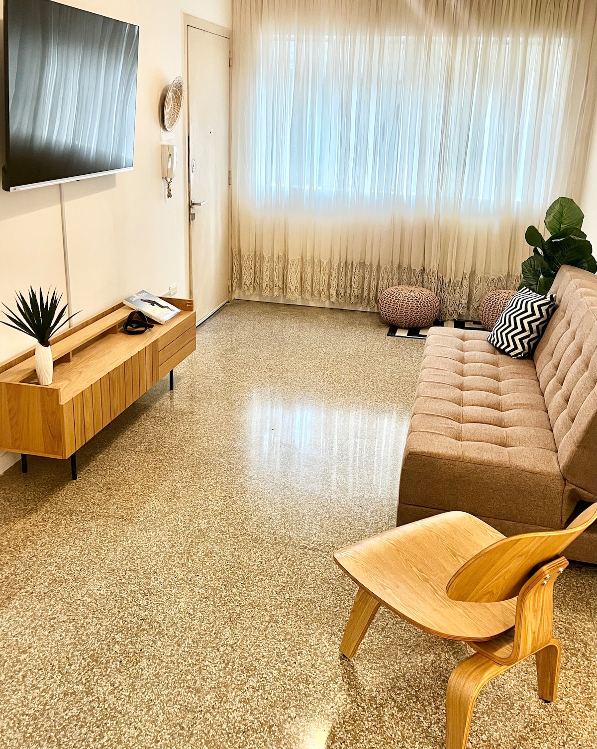 A comfortable living area features a brown sofa bed positioned across from a wall-mounted television. A wooden console table holds decorative items, while natural light filters through sheer curtains. An accent chair and a round pouf add to the inviting layout.