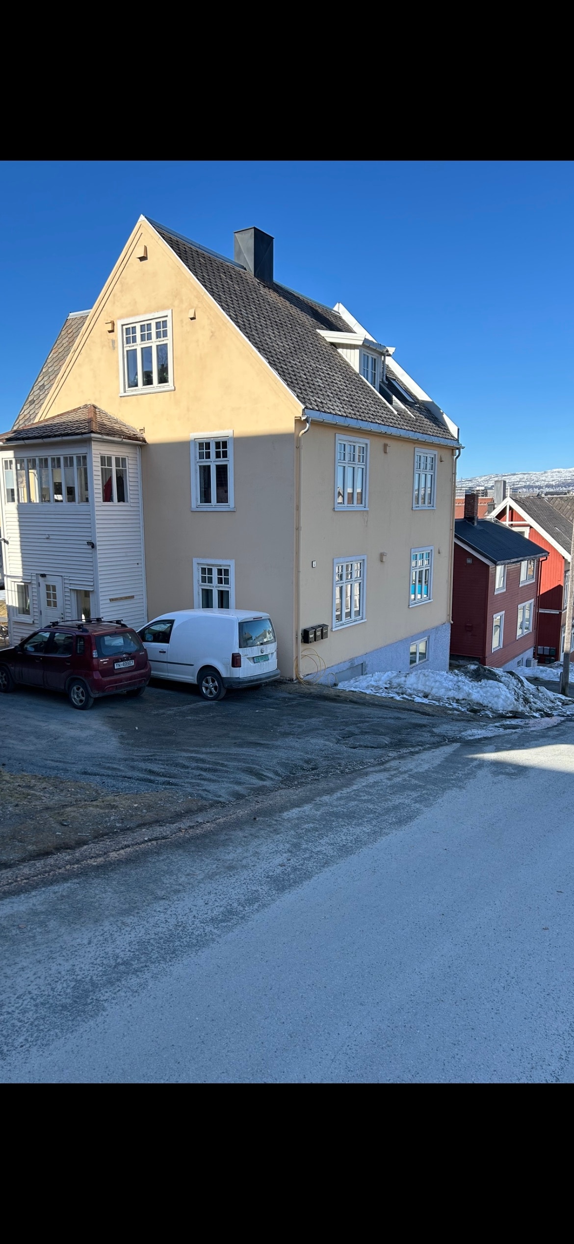 A multi-story building is presented with a light-colored exterior and multiple windows, contrasting with the clear blue sky. Two vehicles are parked in the driveway, and a neighboring red house adds warmth to the scene against the snowy ground.