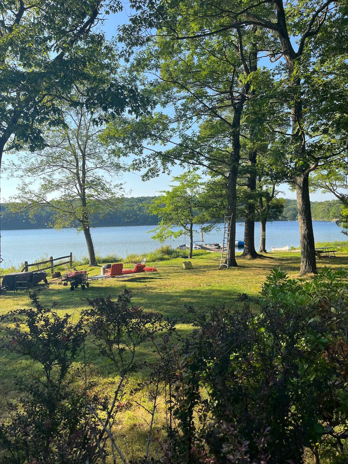 A serene lakeside view is presented, framed by tall trees and lush greenery. Comfortable seating areas with chairs are arranged on the grass, facing the calm water. A distant shoreline is visible, enhancing the sense of tranquility in this natural setting.