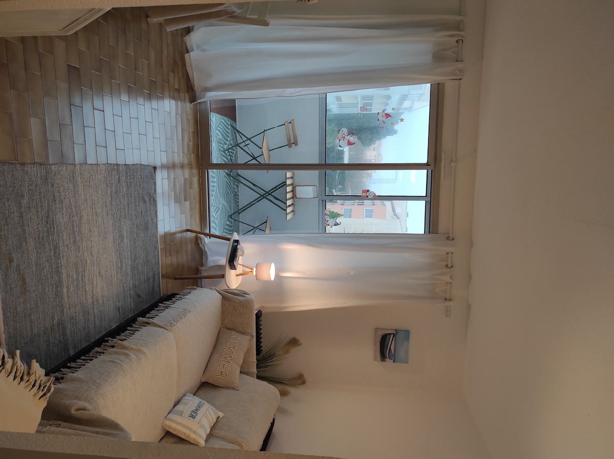 A light-filled seating area is presented, featuring a spacious sofa adorned with neutral-toned cushions. Nearby, a small lamp illuminates a side table. Large glass doors provide views of an outdoor balcony with green folding chairs and a small table, enhancing the room's airy feel.