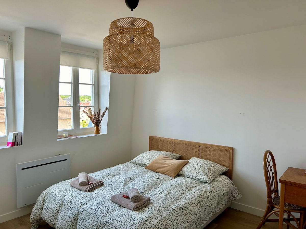 A spacious bedroom is illuminated by natural light streaming through two windows. A queen-size bed is dressed with patterned bedding and complemented by decorative cushions. A wooden desk and a woven pendant light add warmth to the minimalist decor. Soft towels are neatly arranged on the bed.
