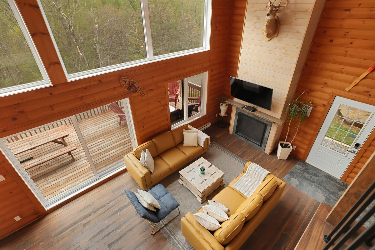A spacious living area showcases large windows that offer views of the surrounding greenery. Cozy seating is arranged around a central coffee table, with a television mounted above a stone fireplace. The wooden walls and flooring create a warm, natural ambiance.