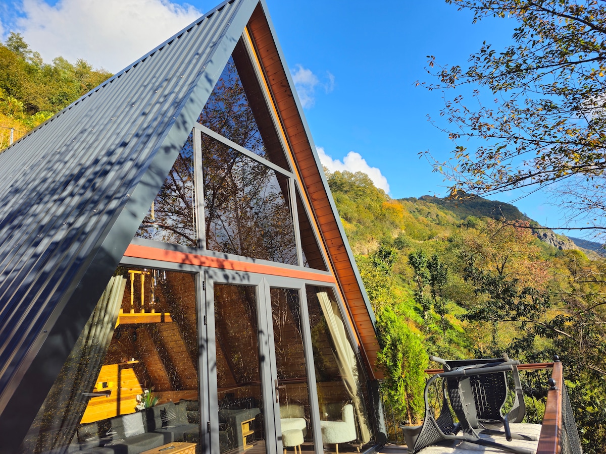 The image showcases a modern A-frame cabin, featuring large glass windows that reflect the surrounding greenery. A wooden deck is visible, accompanied by outdoor seating, inviting relaxation in nature's embrace.