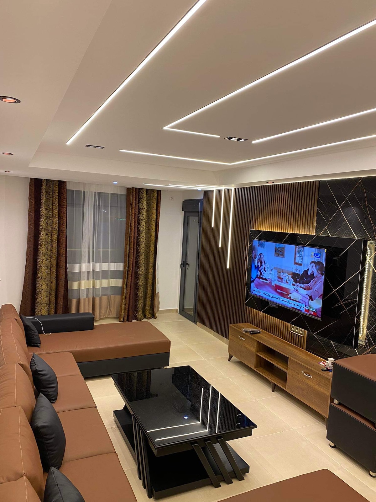 A modern living area features a large sectional sofa in brown tones and a sleek black coffee table. The room is illuminated by recessed lighting, highlighting a wall-mounted TV. Floor-to-ceiling windows are dressed with patterned curtains, creating an inviting atmosphere.