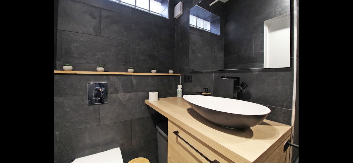 A modern bathroom features dark wall tiles and a light wooden countertop. A round, white vessel sink is positioned on the counter, complemented by a sleek faucet. A shelf holds small decorative items, and natural light filters in through a window above.