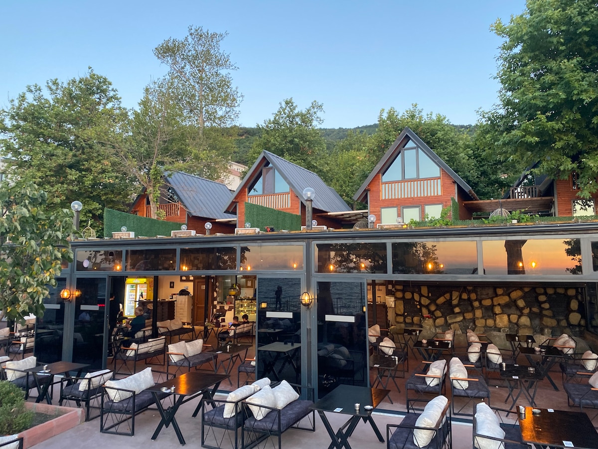 The image displays a welcoming café area featuring dark wooden tables and comfortable seating. Large glass front provides an unobstructed view of the outdoors, showcasing lush greenery and the bungalows in the background. The serene atmosphere is highlighted by gentle lighting fixtures.