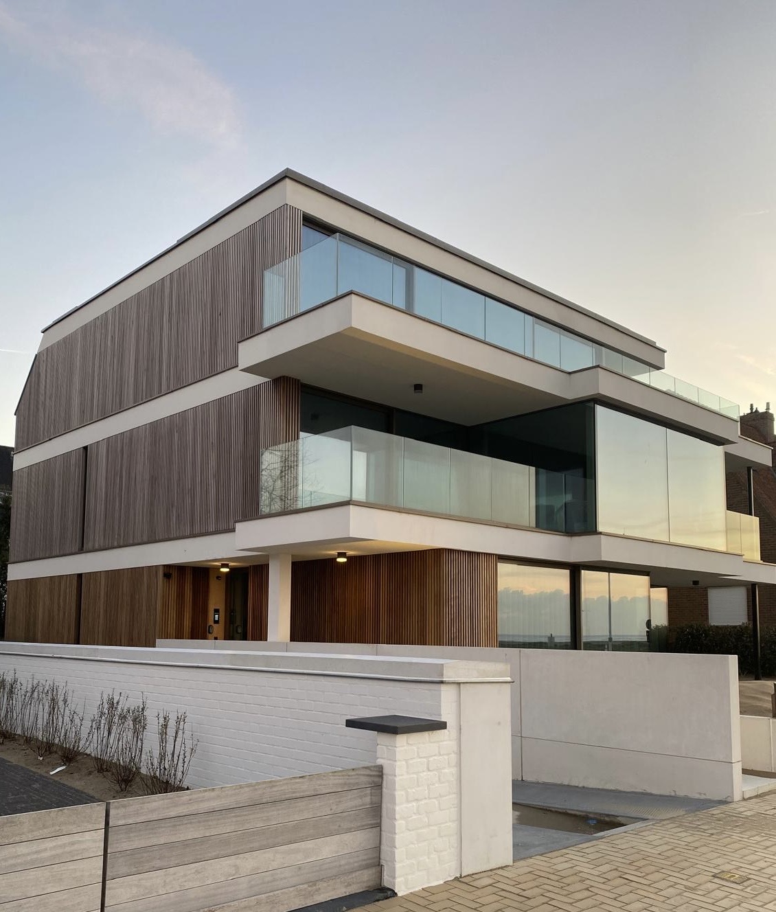 The modern three-story building features wooden and glass facades, creating an elegant exterior. Balconies are visible on each level, offering views of the surroundings. A minimalist front gate leads to a well-maintained pathway, emphasizing the contemporary architectural style.