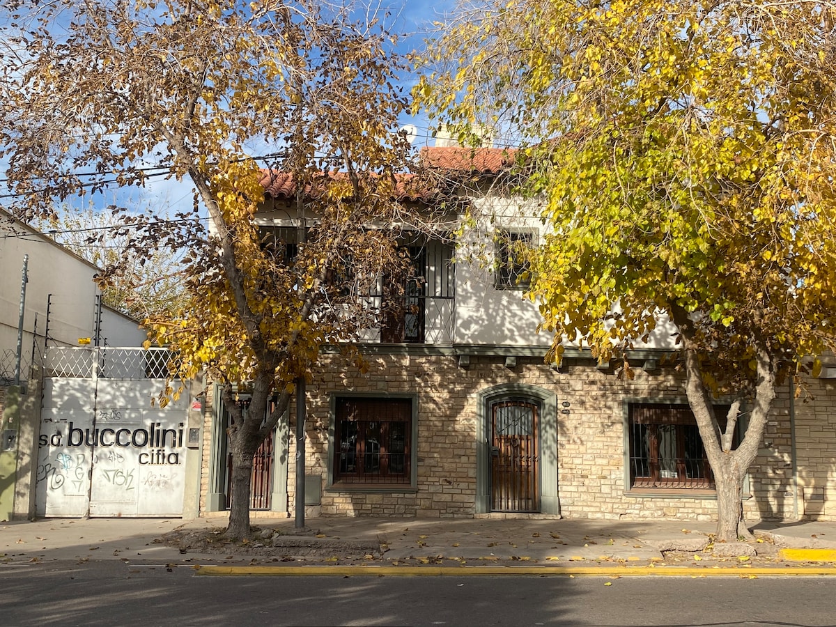 A historic two-story chalet with a stone exterior is framed by vibrant autumn foliage. Large windows are adorned with wrought iron, allowing natural light to filter through. The entrance features a wooden door and a gated pathway, creating an inviting ambiance.