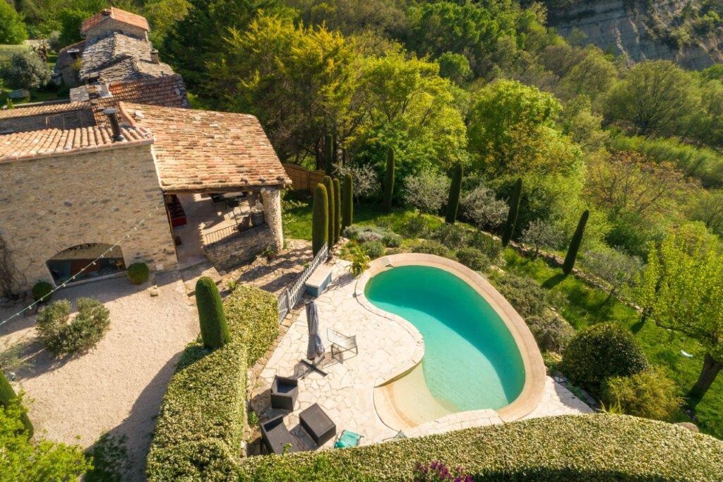 An aerial view showcases a serene swimming pool surrounded by lush greenery and neatly trimmed hedges. A patio area with sun loungers and a small outdoor lounge is visible, highlighting the inviting outdoor space.  The stone house blends harmoniously with the natural landscape.