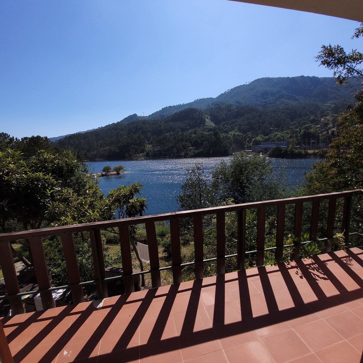 A wide balcony provides a view of the shimmering lake bordered by lush greenery and distant mountains. The sun casts a gentle glow on the water, creating a serene atmosphere. The wooden railing frames the picturesque scene, emphasizing the connection to nature.